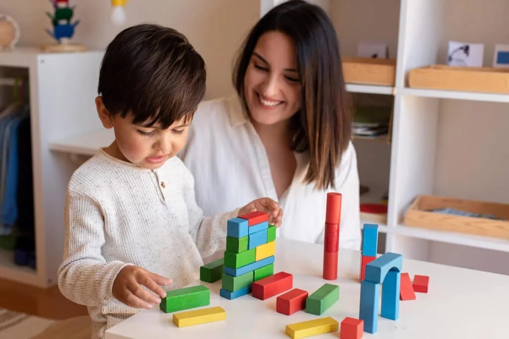Liltle Preschooler Kid playing with wood blocks and teacher educador help. Homeshooling. Learning Community. Montessori School