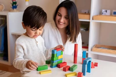 Liltle Preschooler Kid playing with wood blocks and teacher educador help. Homeshooling. Learning Community. Montessori School